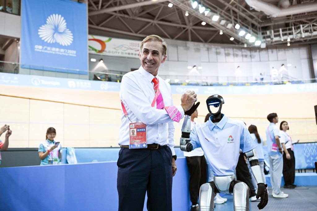 11月13日，一名賽事技術官員在場地自行車女子團體競速賽決賽前同參與表演的機器人擊掌合影。