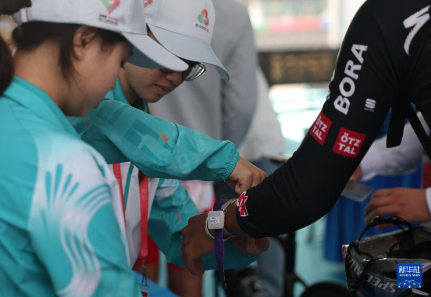 公路自行車賽道上的口岸區域安裝了無線射頻識別系統（RFID）。這套系統能在選手經過時自動讀取車手與車的電子芯片，并即時回傳到后臺邊檢、海關系統進行同步核驗，形成全程閉環管理。圖為志愿者為公路自行車男子個人賽的參賽選手佩戴通關手環。